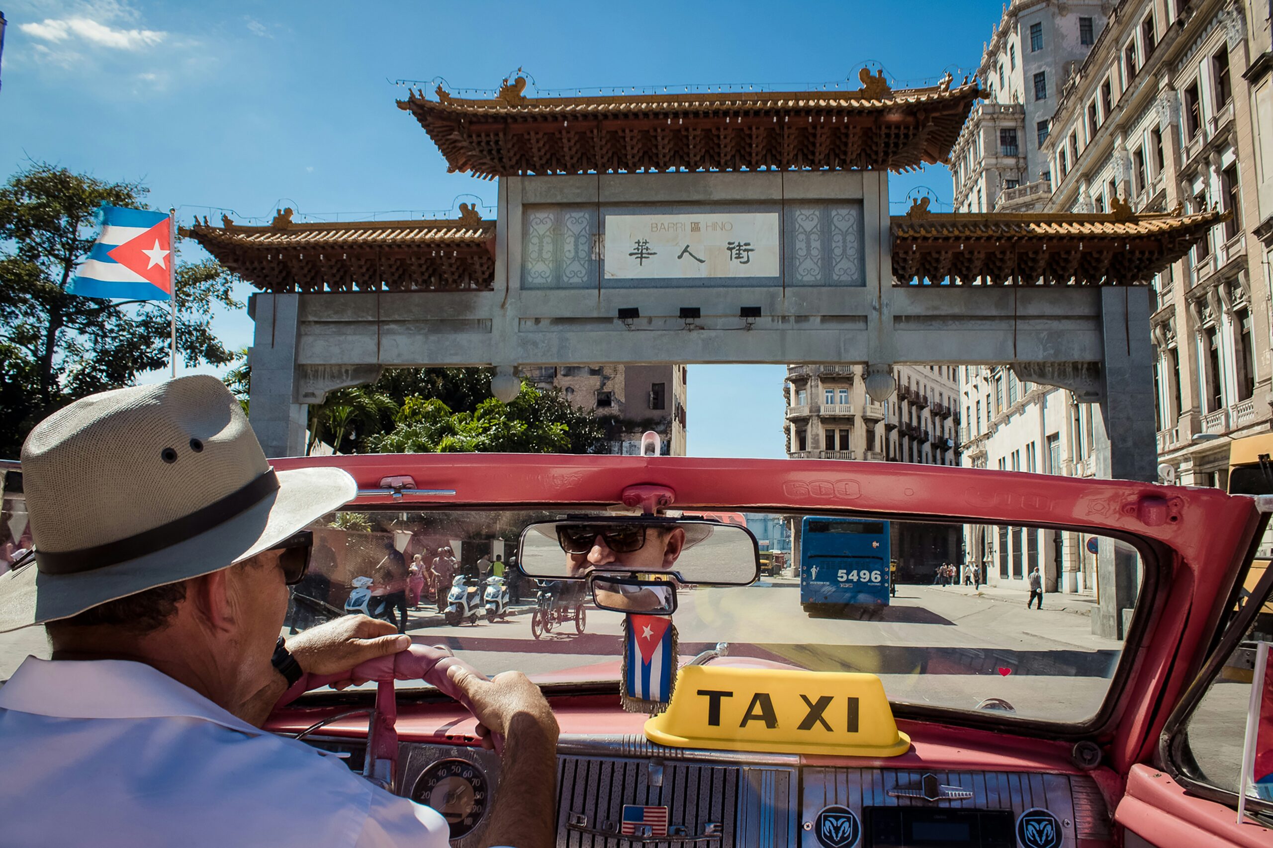 A man drives a classic convertible taxi with a "TAXI" sign past Havana's Chinatown gate, while a Cuban flag is visible on the left.