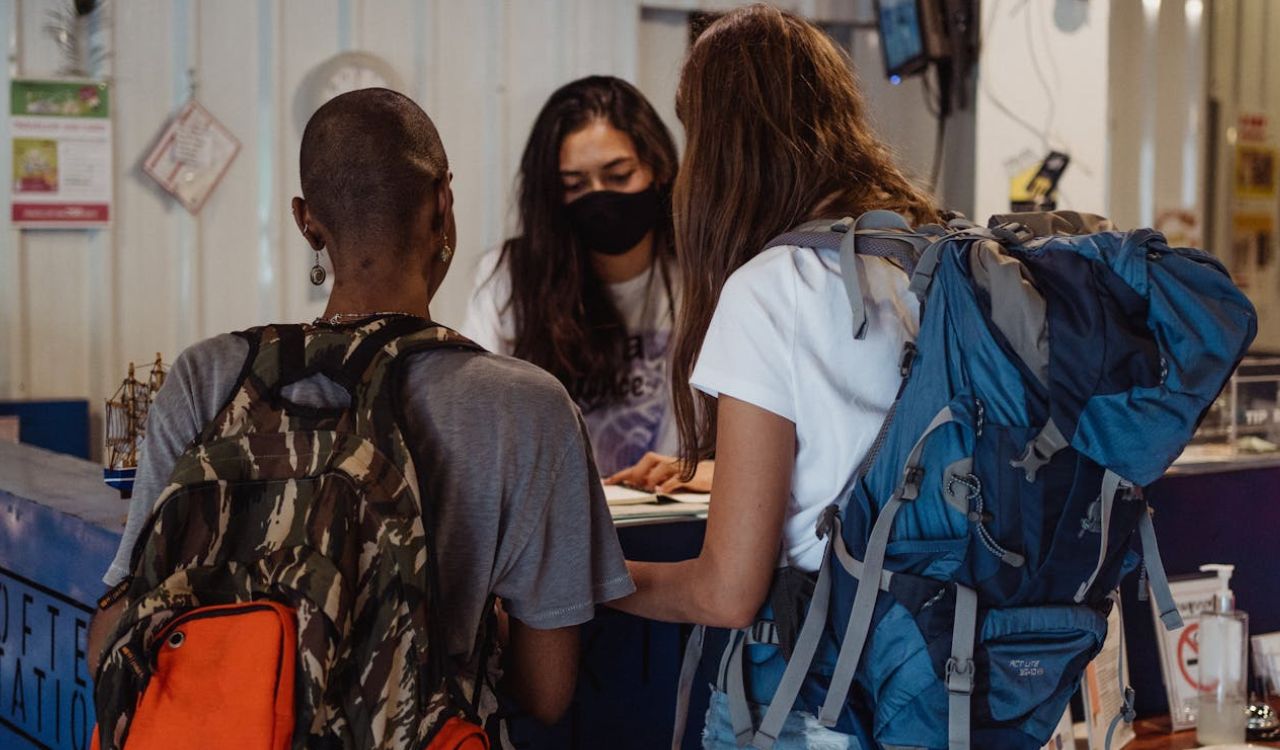 Two backpackers checking in at a reception desk with a staff member wearing a mask