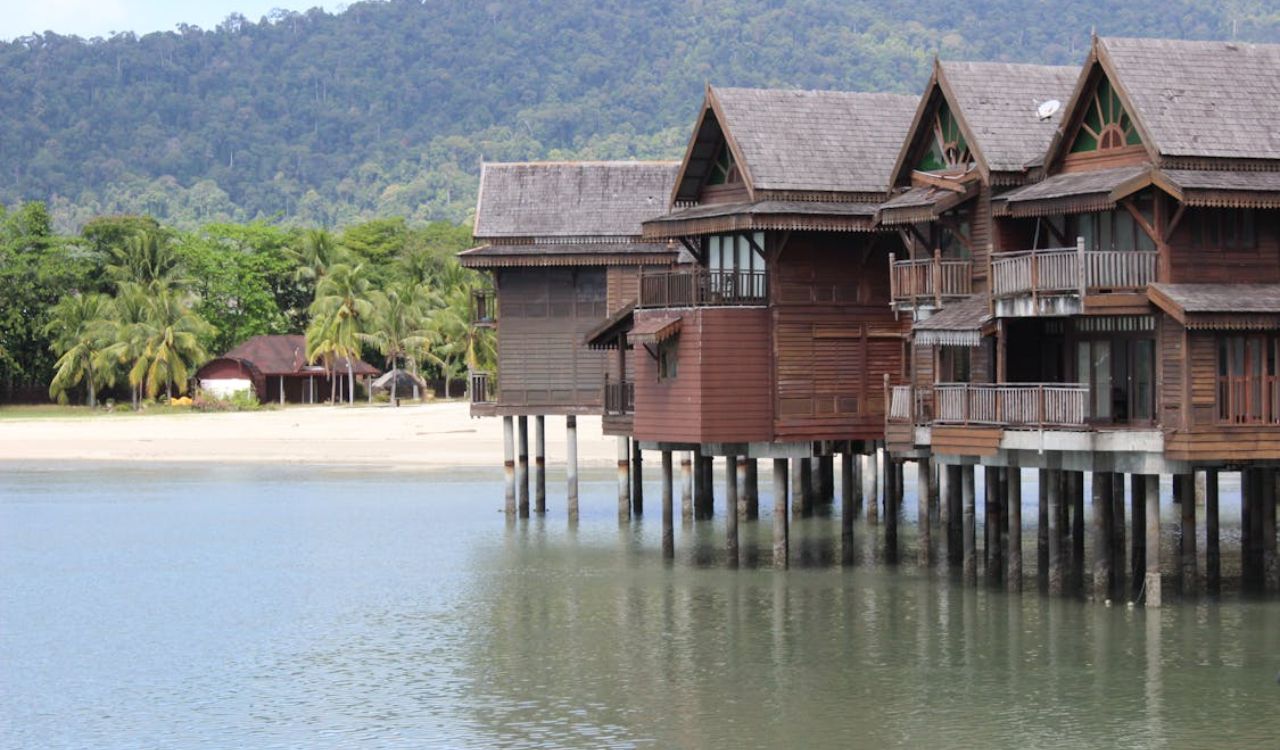 Wooden beachfront houses on stilts over calm water, with a sandy beach and palm trees in the background