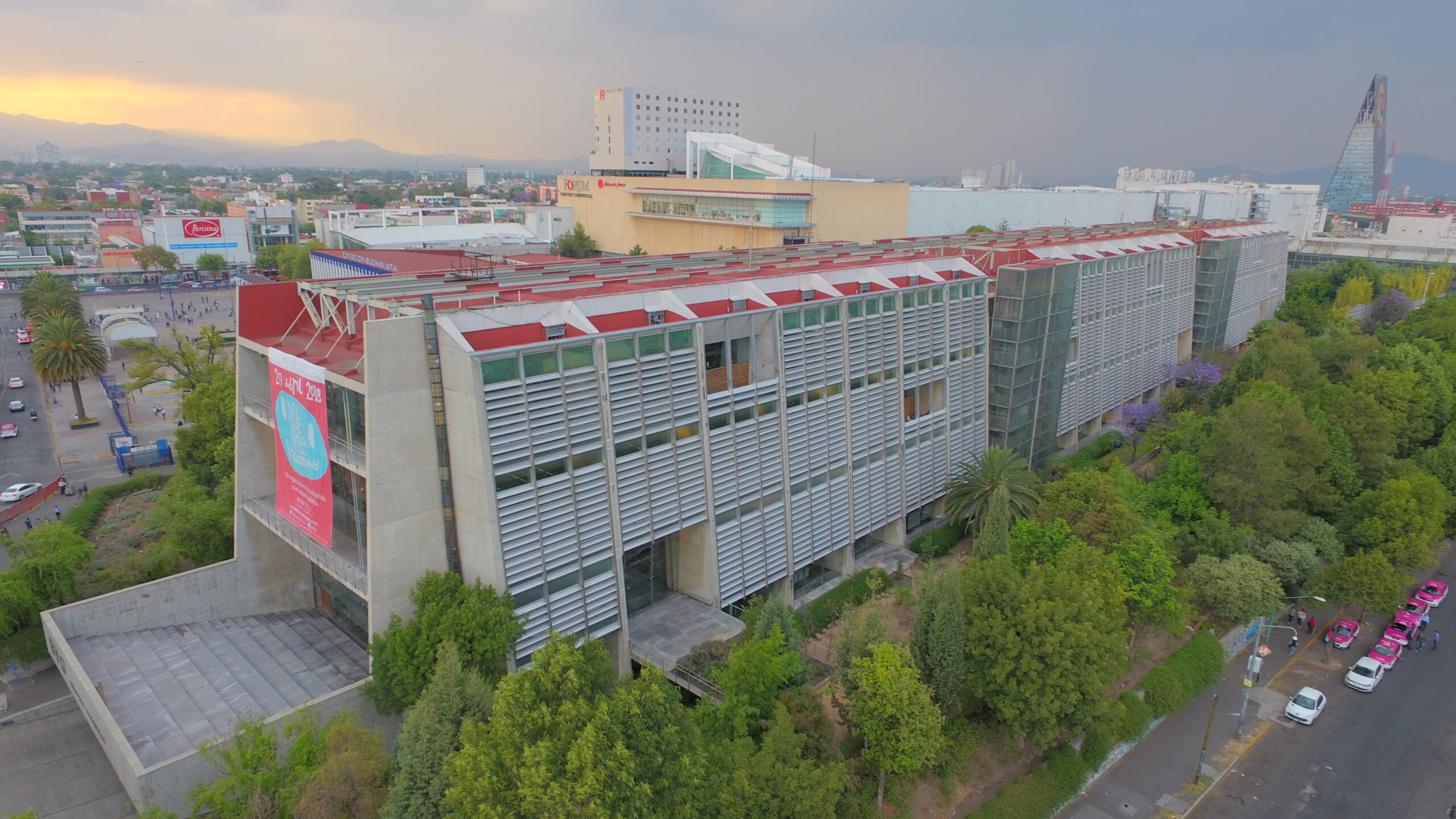 A large modern building with horizontal metal slats and a red roof stands among trees, with a cityscape in the background beneath a cloudy sky.
