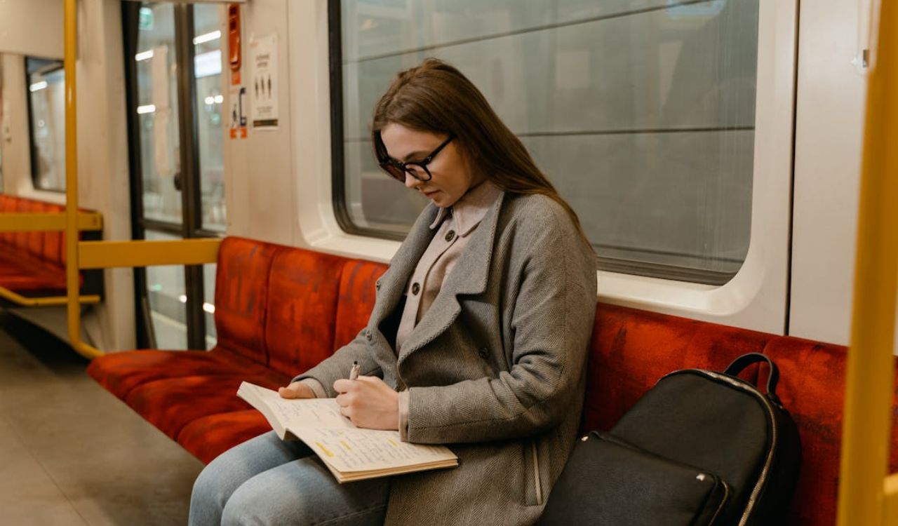 Woman writing in notebook while sitting on a subway train with a backpack beside her