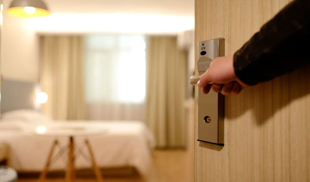 Hand opening a hotel room door with a cozy, well-lit interior visible inside