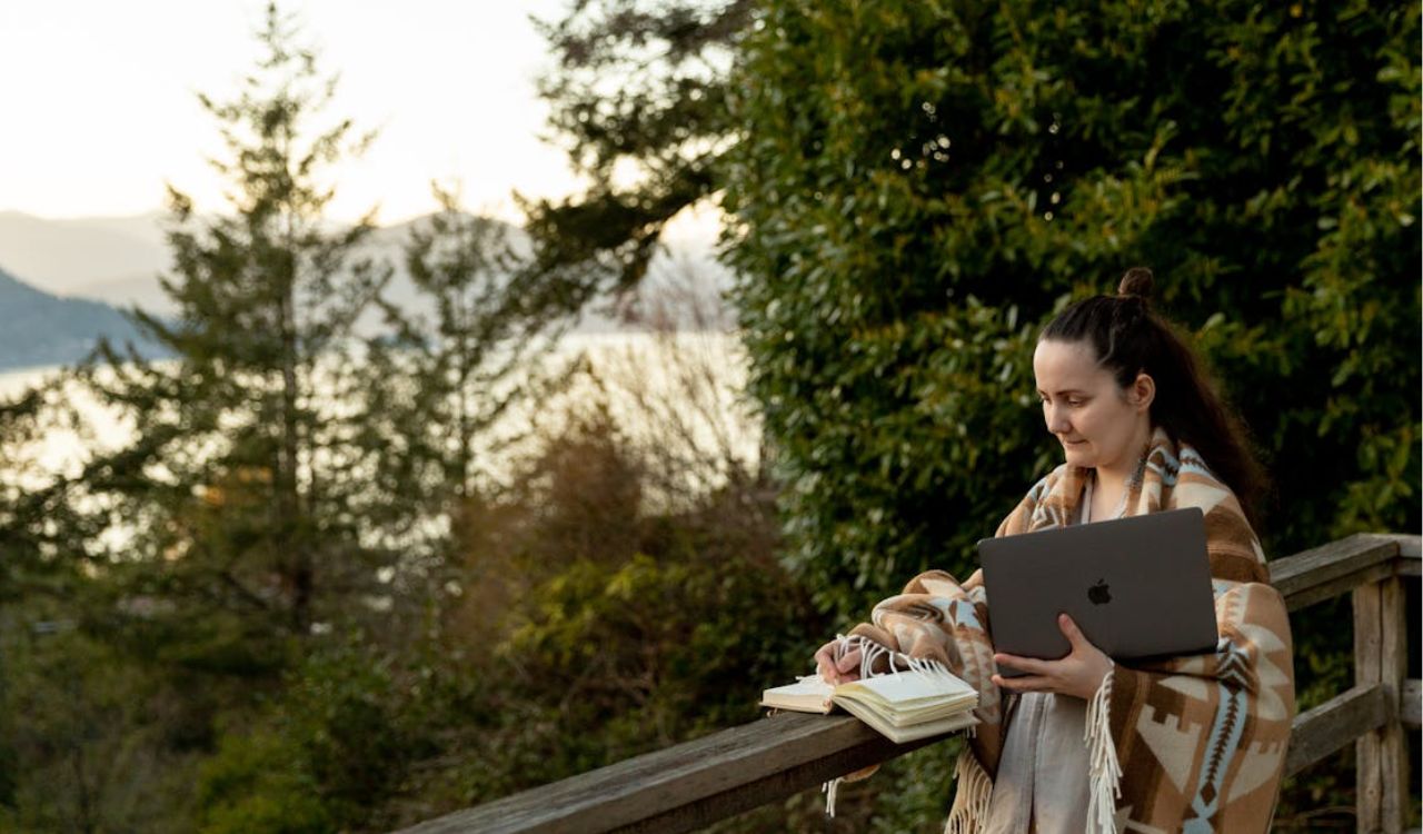 Woman working on a laptop while holding a notebook outdoors, wrapped in a cozy blanket with nature in the background