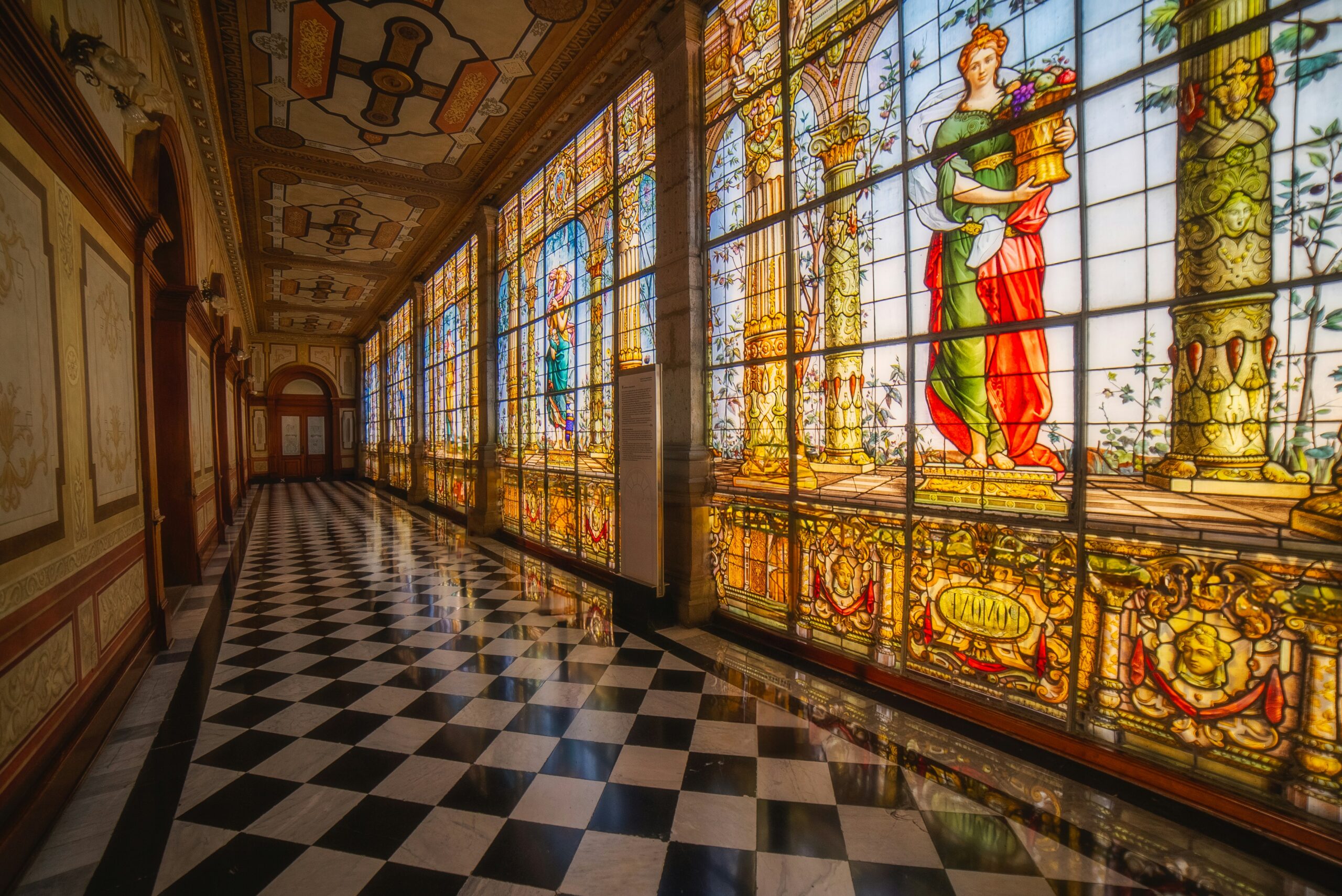 Ornate hallway with a black-and-white checkered floor, large stained glass windows of classical figures, and decorative columns.