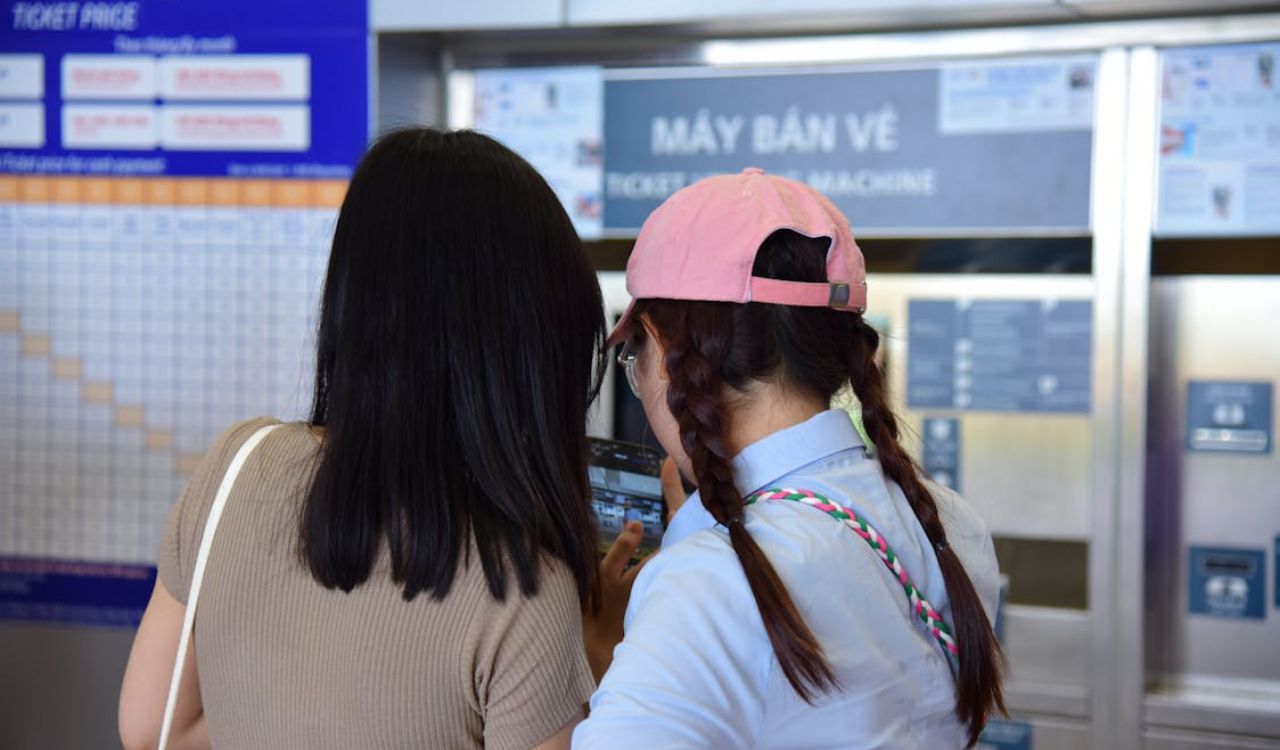Two people standing in front of a ticket machine, reviewing information displayed in a station with a price chart in the background