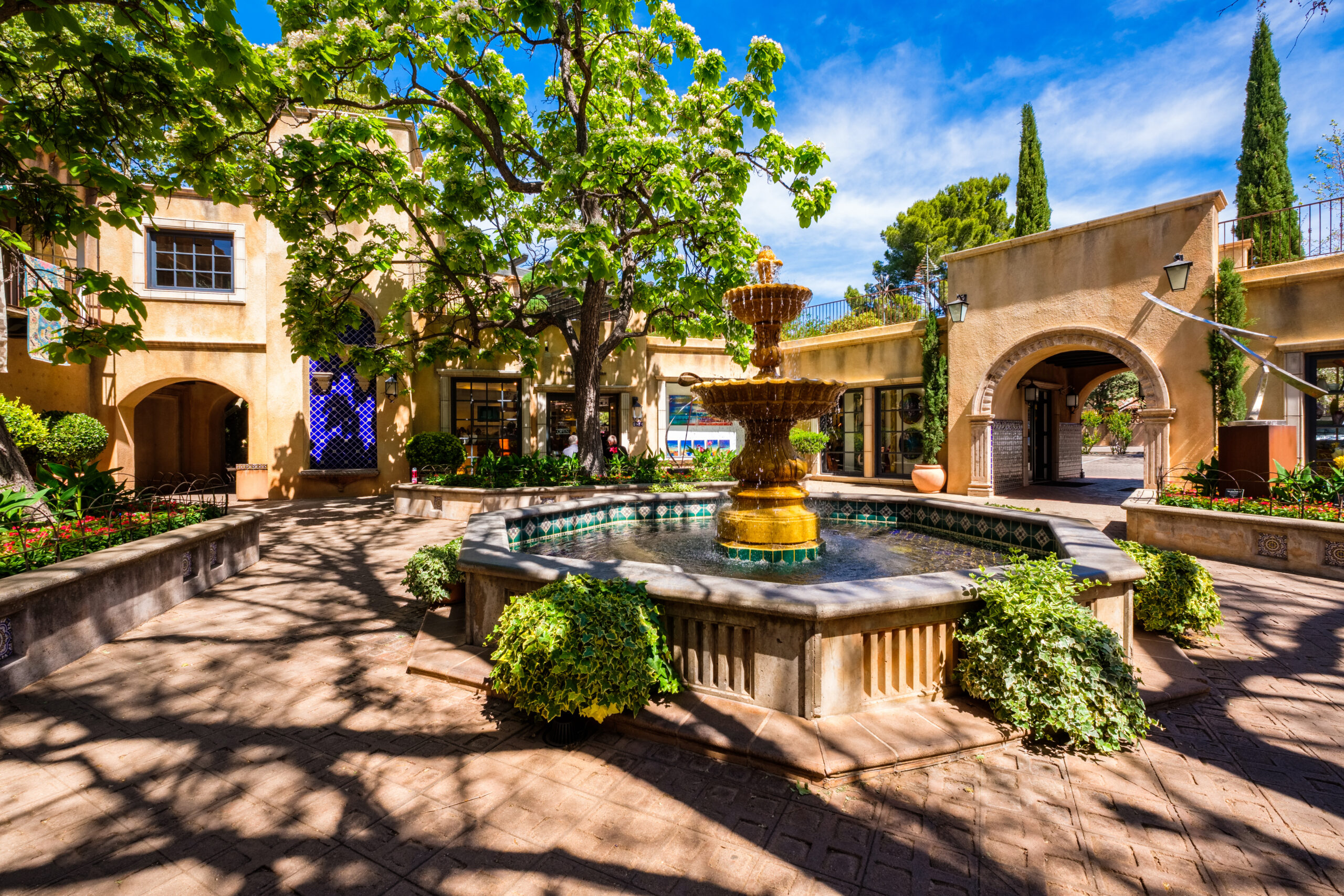 A central tiered fountain stands in a courtyard surrounded by greenery, trees, and stucco buildings beneath a clear blue sky.