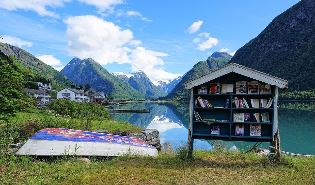 Outdoor bookshelf in Fjærland’s Book Town beside mirror-calm fjord and snow-capped Norwegian mountains
