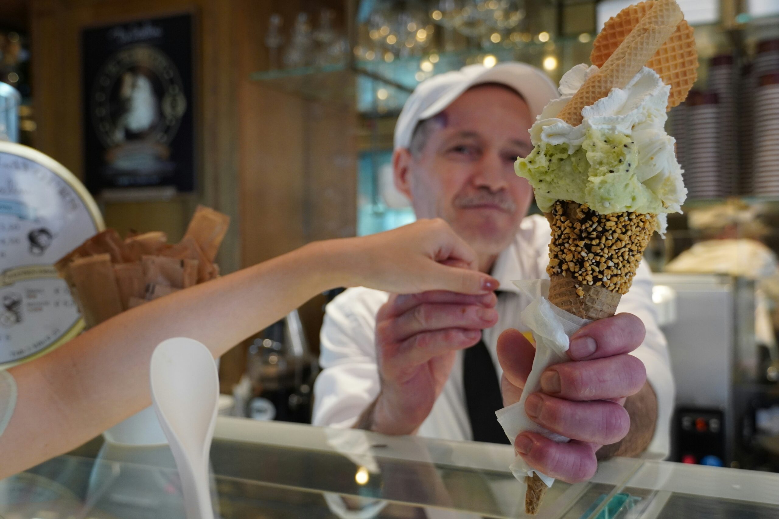 A person hands a pistachio and vanilla ice cream cone with wafers and sprinkles to a customer across the counter.
