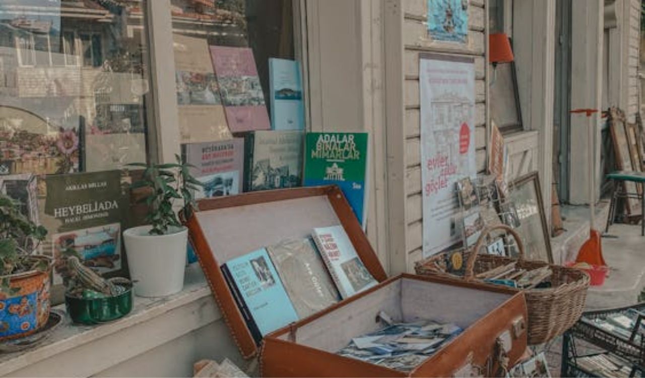 Outdoor display of books, vintage magazines, and souvenirs in front of a shop window, with a suitcase and baskets on the ground