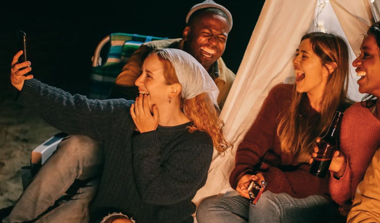 Friends laughing and taking a selfie around a campfire at night.