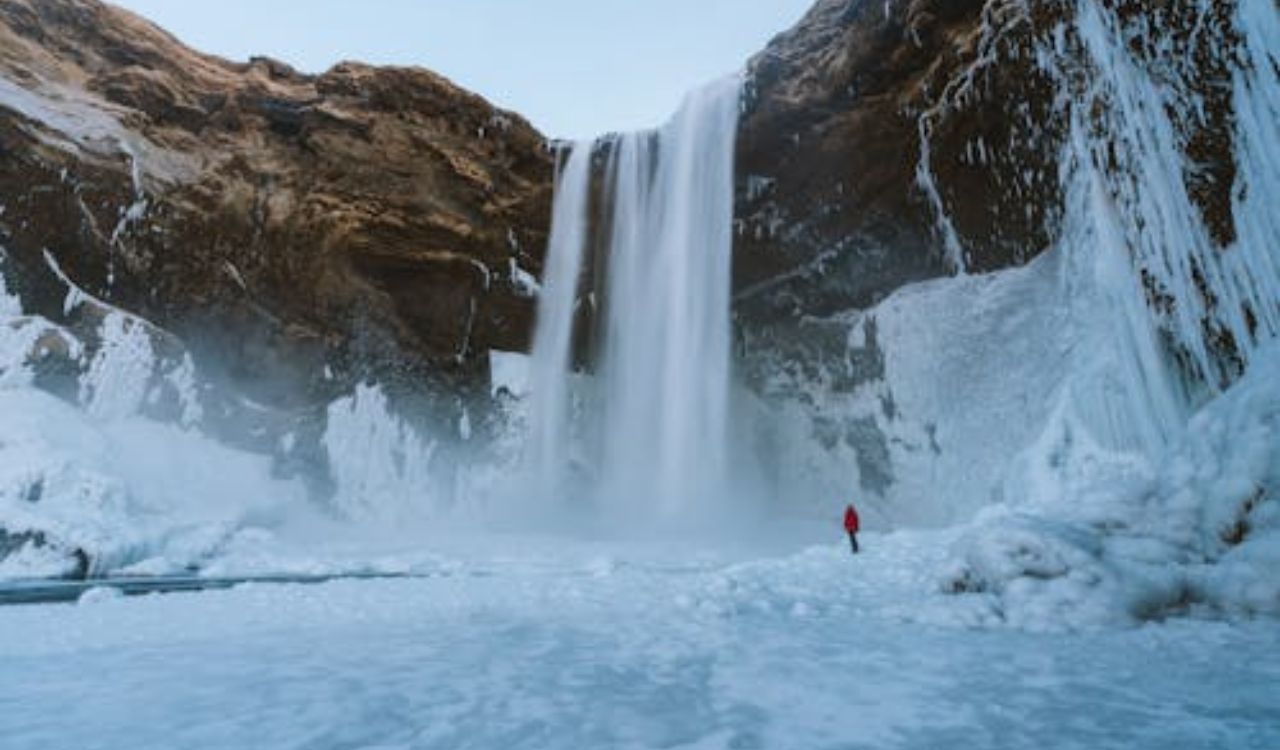 Frozen Icelandic waterfall with snow-covered cliffs and a lone traveler in red coat