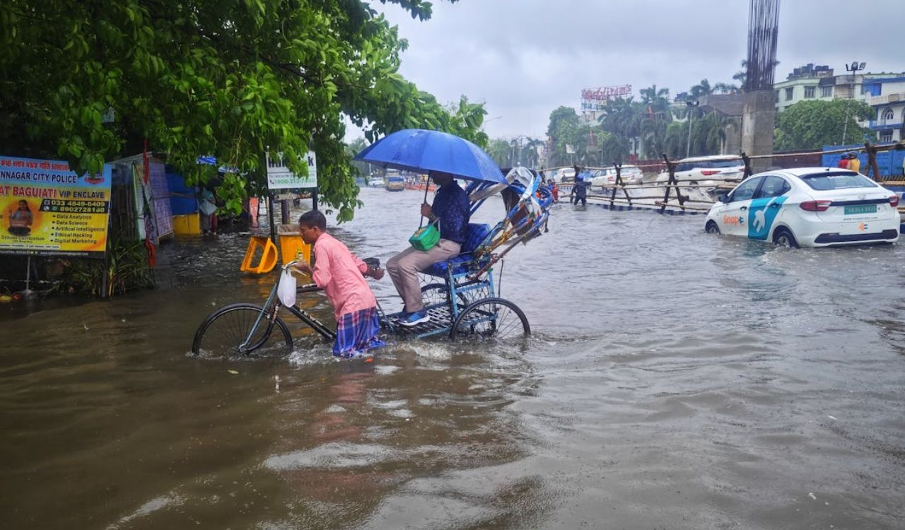 Man pushes a rickshaw through a heavily flooded street during rain, while the passenger holds an umbrella