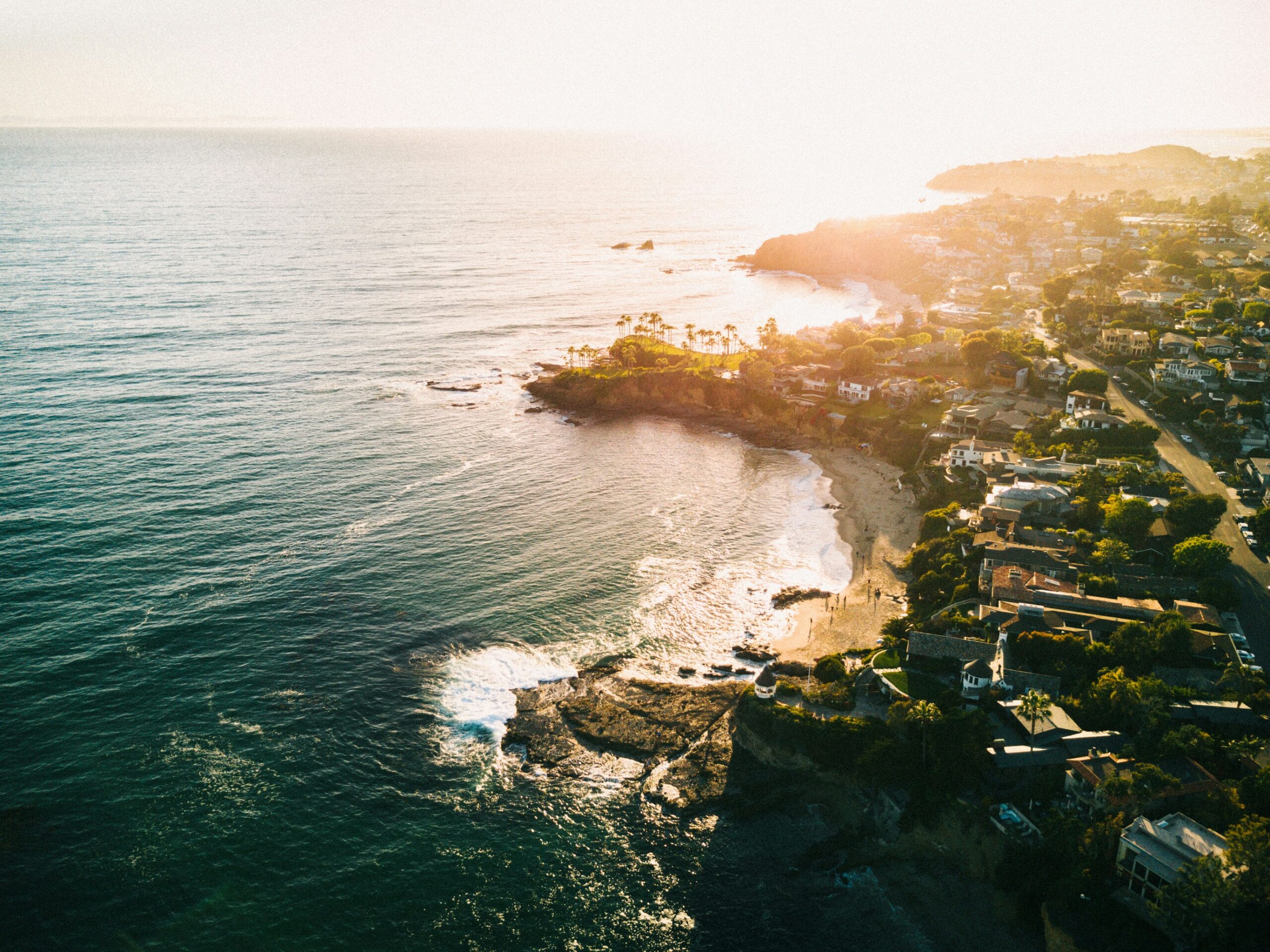 Aerial view of a coastal town at sunset with houses lining the shore and waves crashing on rocky beaches.