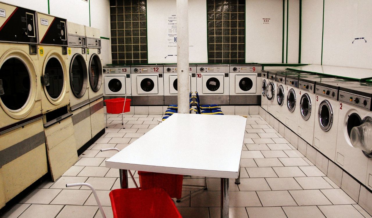 Empty laundromat with washing machines, dryers, red chairs, and a white table.