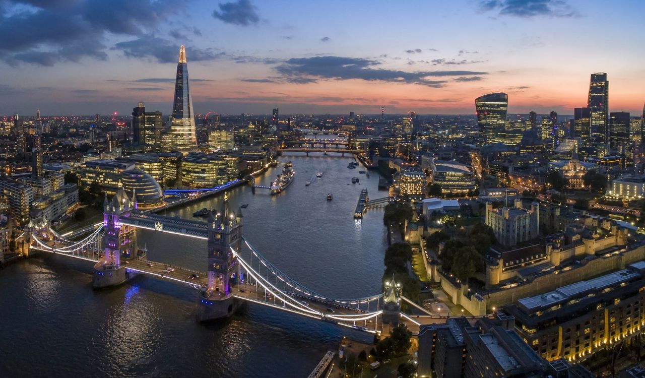 A stunning aerial view of London at twilight, featuring the Tower Bridge, the Thames River, and iconic skyscrapers like The Shard and the Gherkin