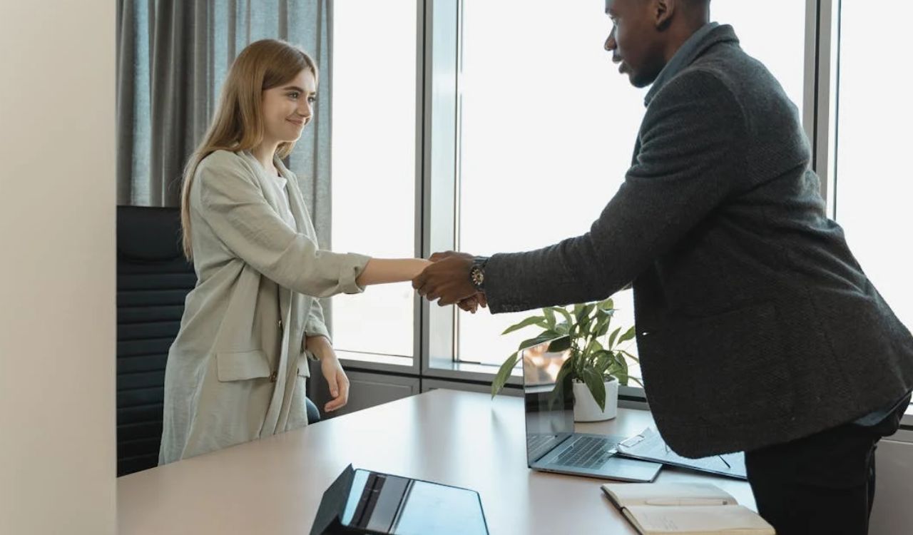 Two professionals shaking hands across a desk in a modern office.