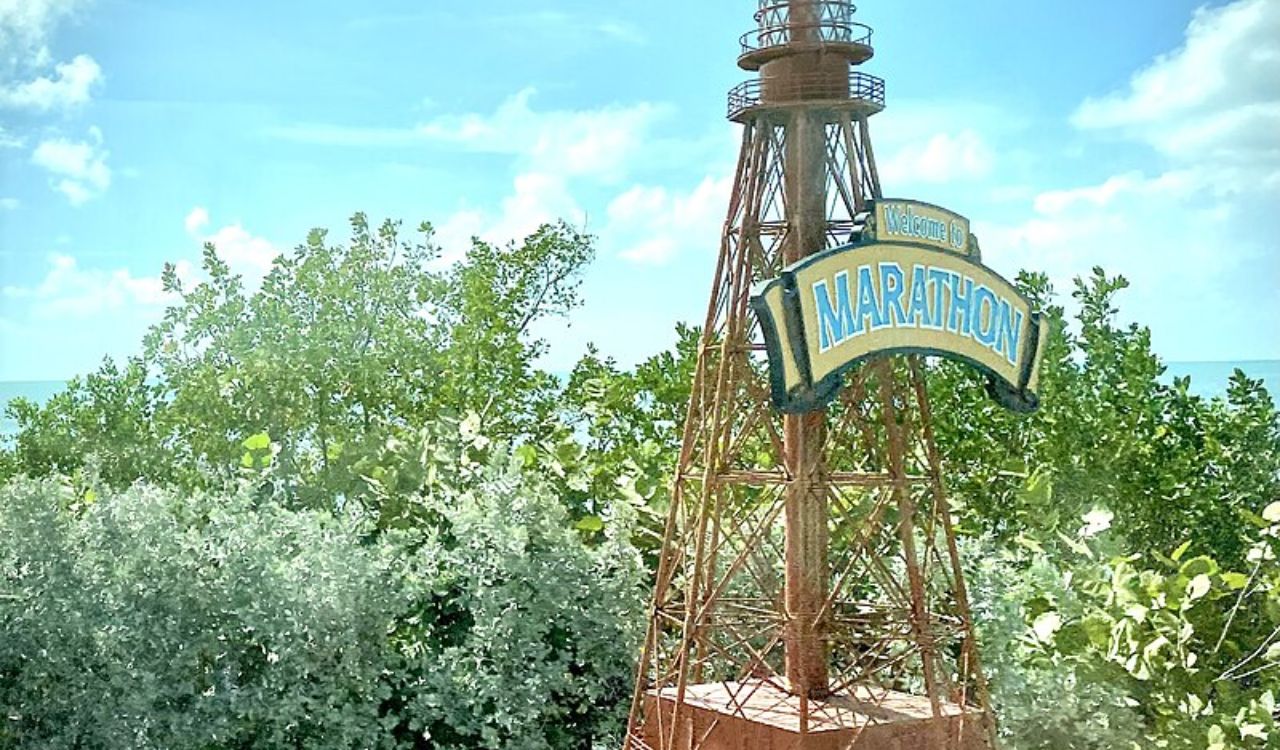 A welcome sign for Marathon, Florida, mounted on a rustic metal tower surrounded by lush green foliage under a bright blue sky