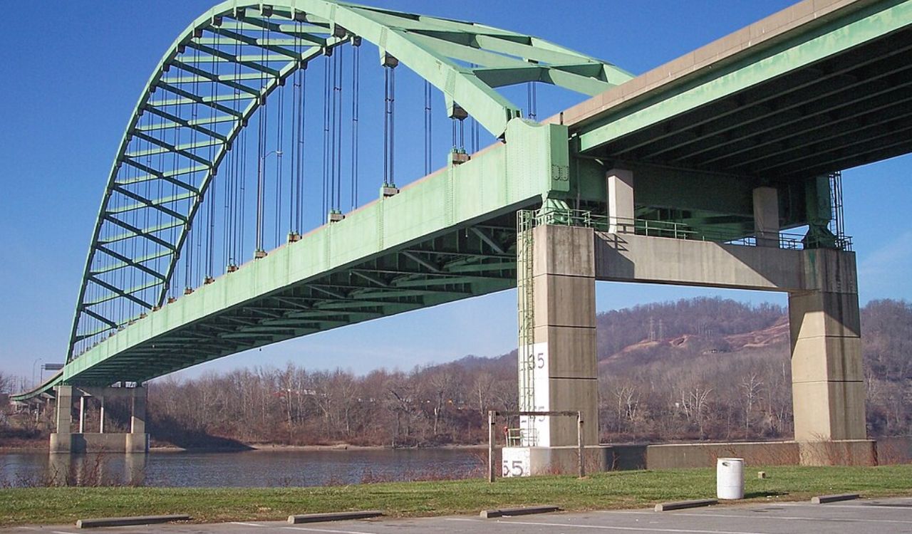 A green suspension bridge over a river with concrete pillars.