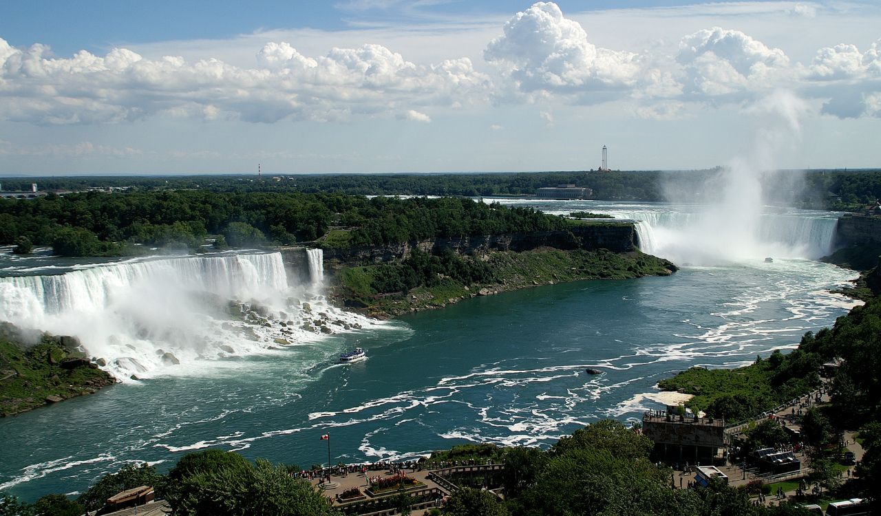 Niagara Falls with mist rising, tour boat below, and lush greenery around.