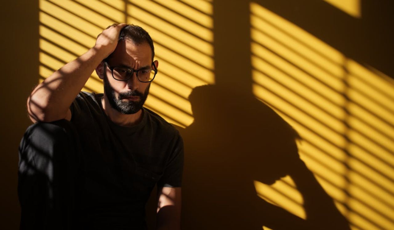 Man sitting alone, looking stressed, with dramatic shadow lines cast by window blinds