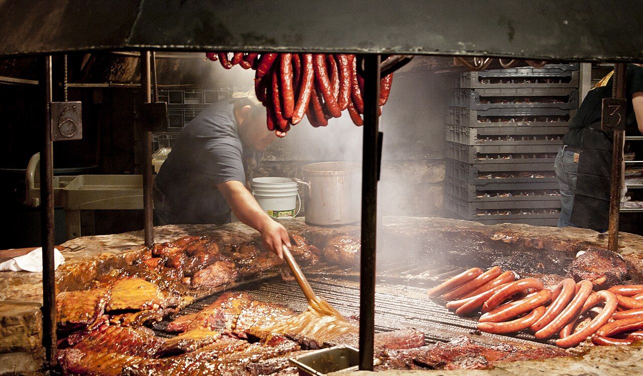 A pitmaster brushes sauce over sizzling brisket and sausages at a smoky Texas barbecue joint