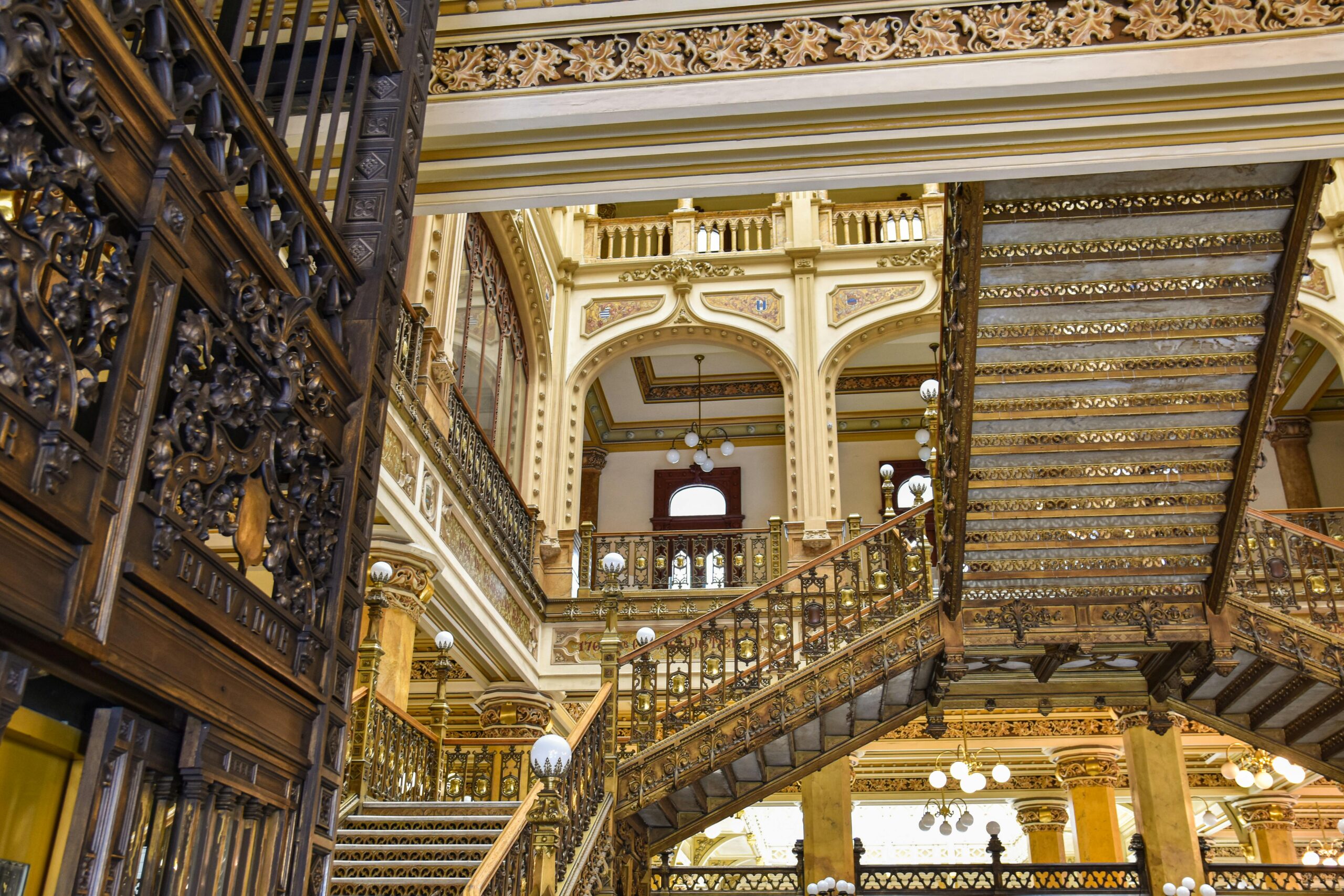 Ornate historic interior featuring decorative metal railings, detailed columns, and multiple staircases under warm lighting.
