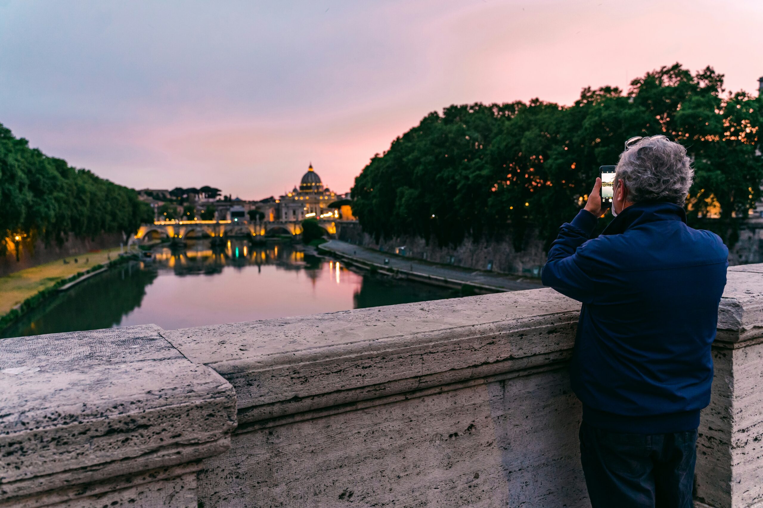 At sunset in Rome, a person stands on a stone bridge photographing St. Peter's Basilica and the Tiber River lined with trees.