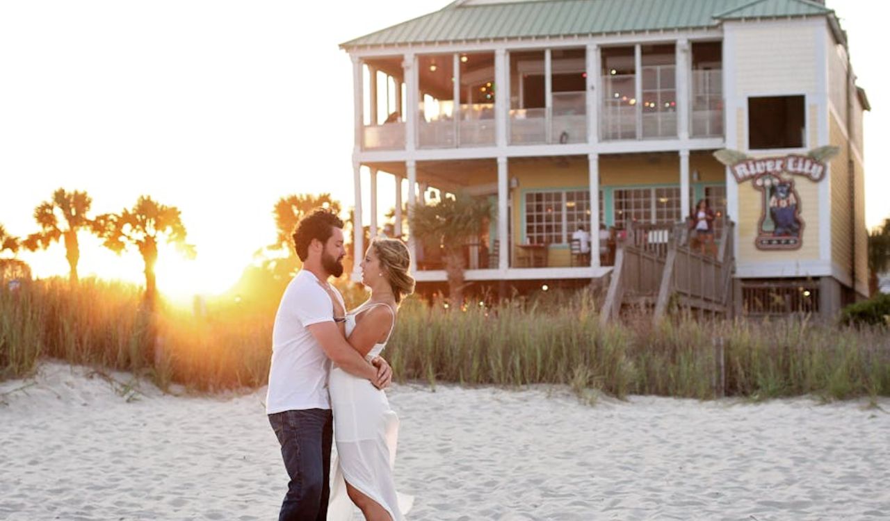 A couple embraces on a sandy beach at sunset near a beach house