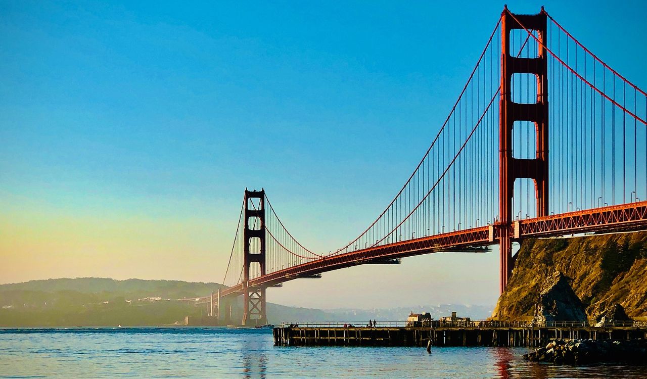 Golden Gate Bridge with a clear blue sky, towering over the water with a rocky shoreline below and a dock extending into the water, captured during daylight