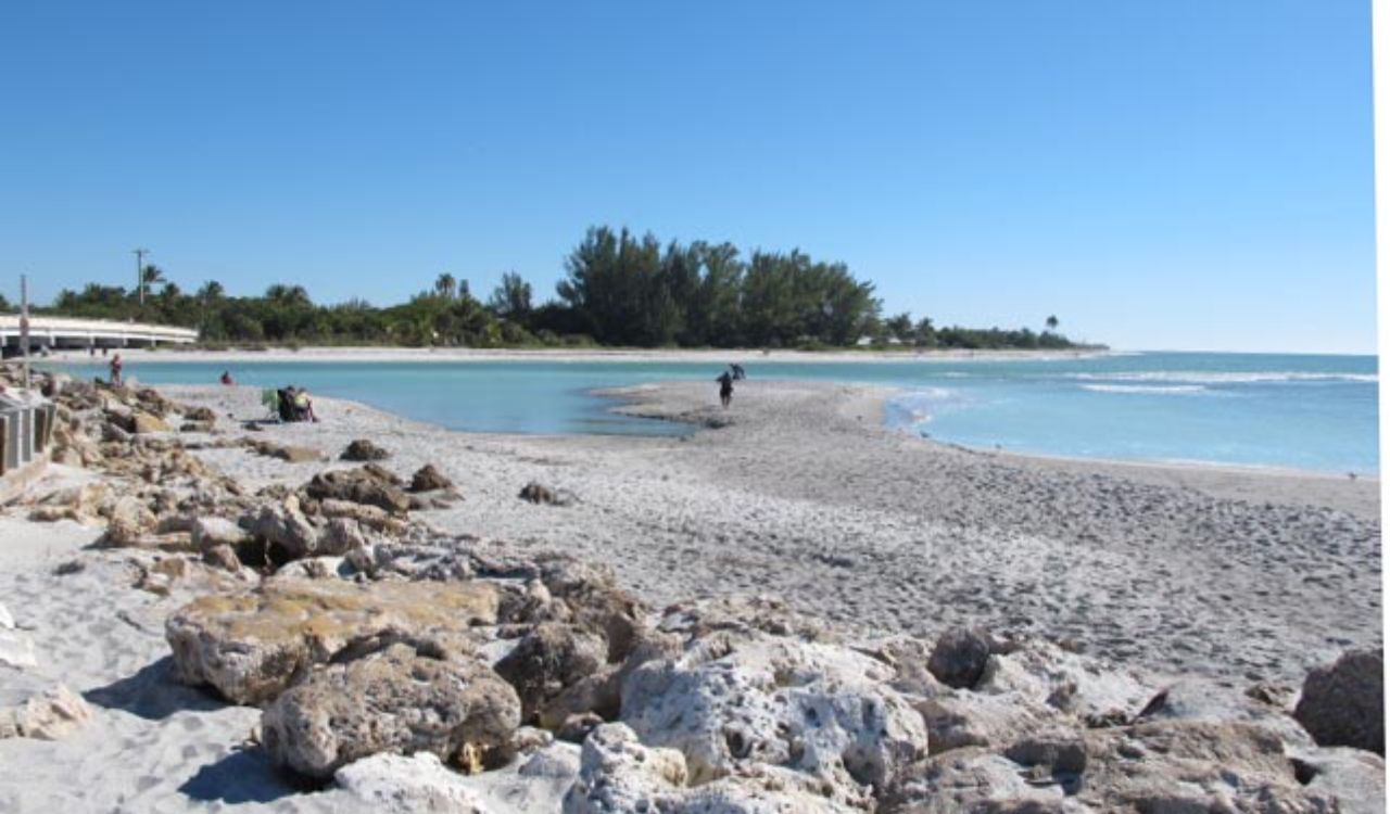 Person walking along sandy beach on Sanibel Island, Florida, with clear blue water and rocky shoreline