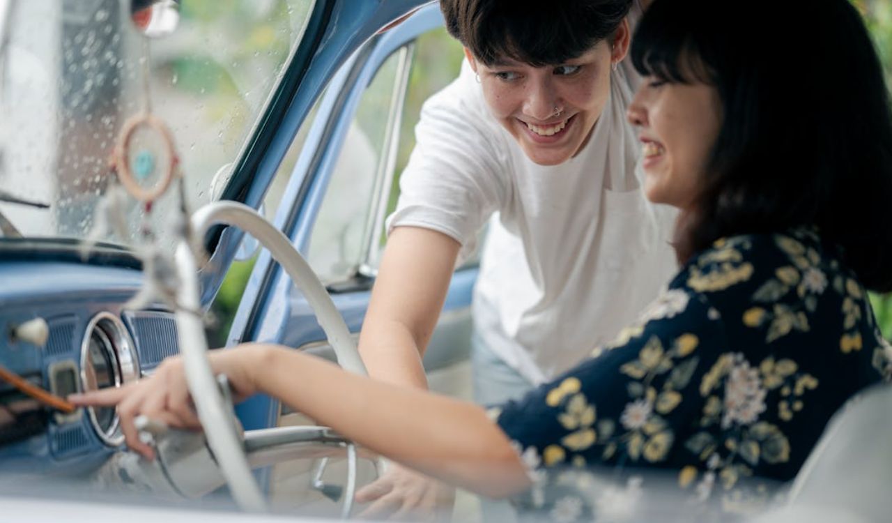 Smiling couple inside a vintage car, with one person pointing at the dashboard and the other leaning in from the driver's side ideal for shared driving travel tips.
