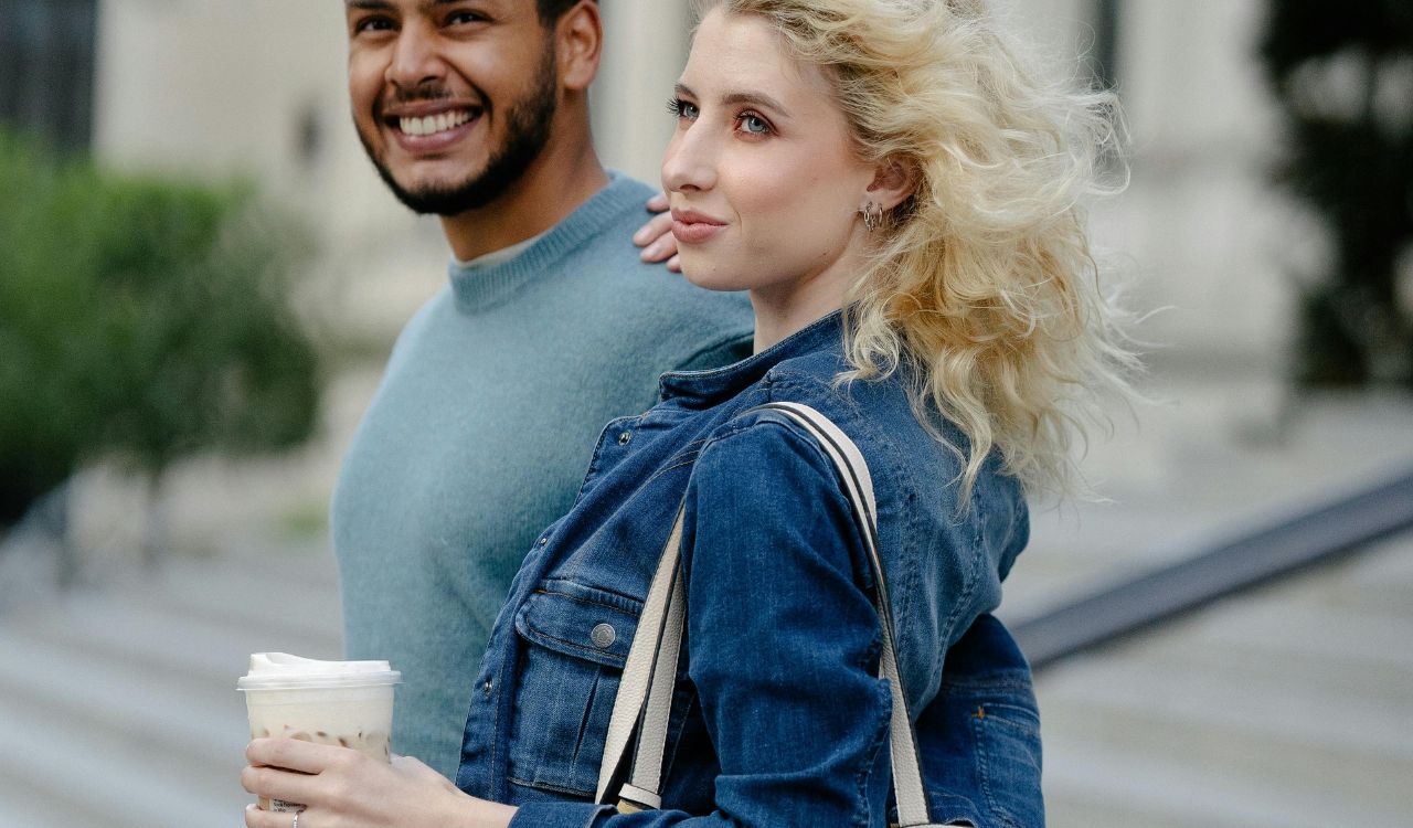 Smiling couple walking outdoors, woman holding a drink and tote bag.