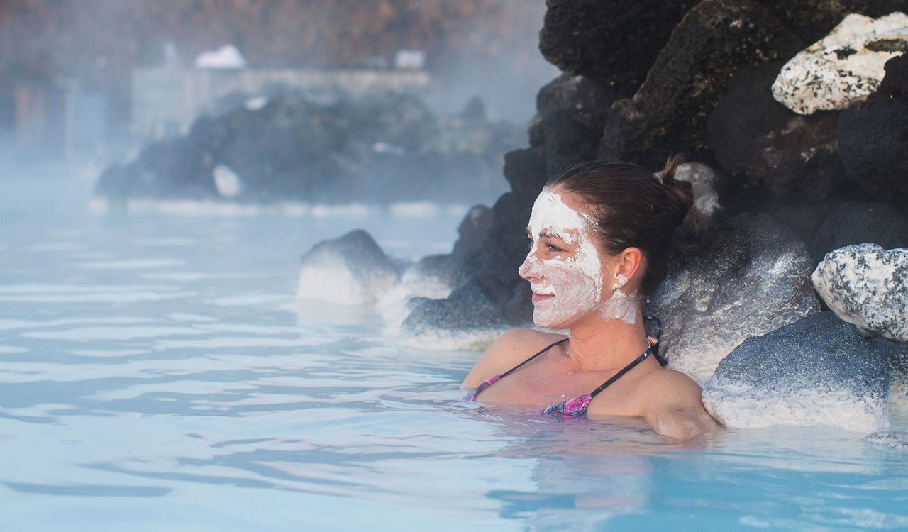Woman relaxing in the Blue Lagoon geothermal spa in Iceland, with a silica mud mask on her face, surrounded by steam and volcanic rocks