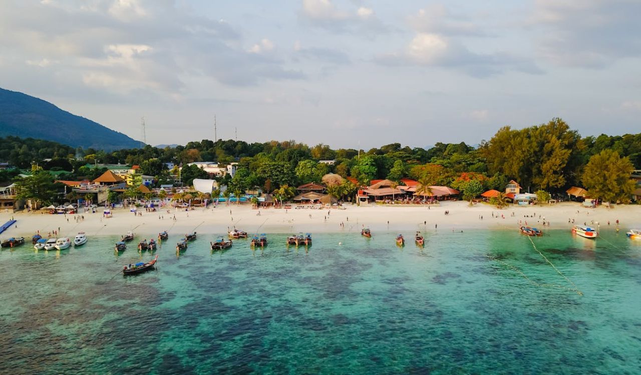 Colorful boats float off Thailand’s white-sand beach, backed by lush greenery and lively seaside buildings