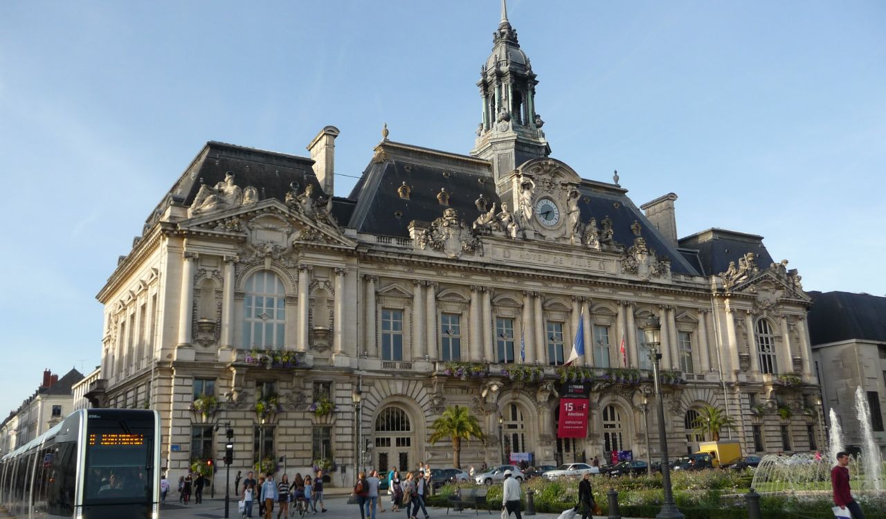 Historic European building with clock tower, tram, and people walking nearby.