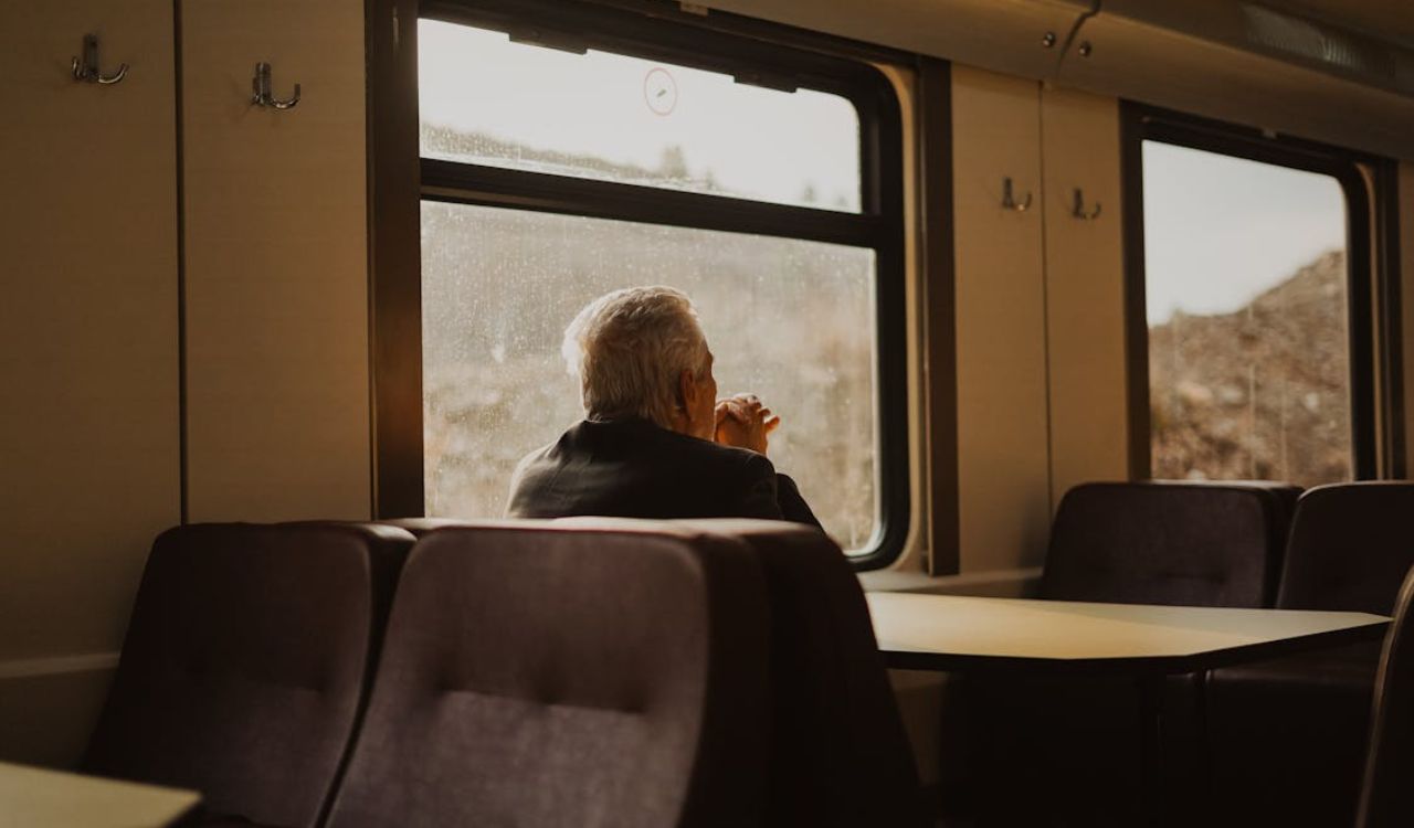 Older man sitting alone by the window in a train dining car, looking out thoughtfully
