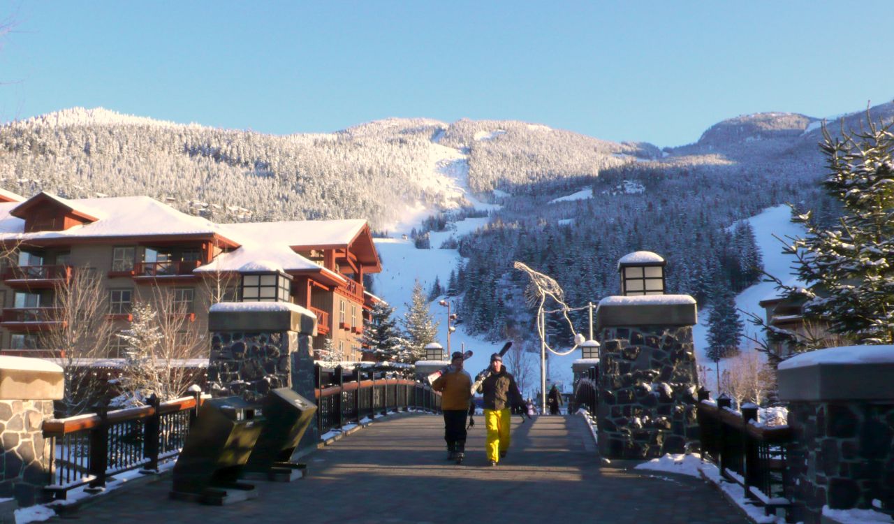Skiers walk across a snowy bridge in Whistler with ski slopes in the background.