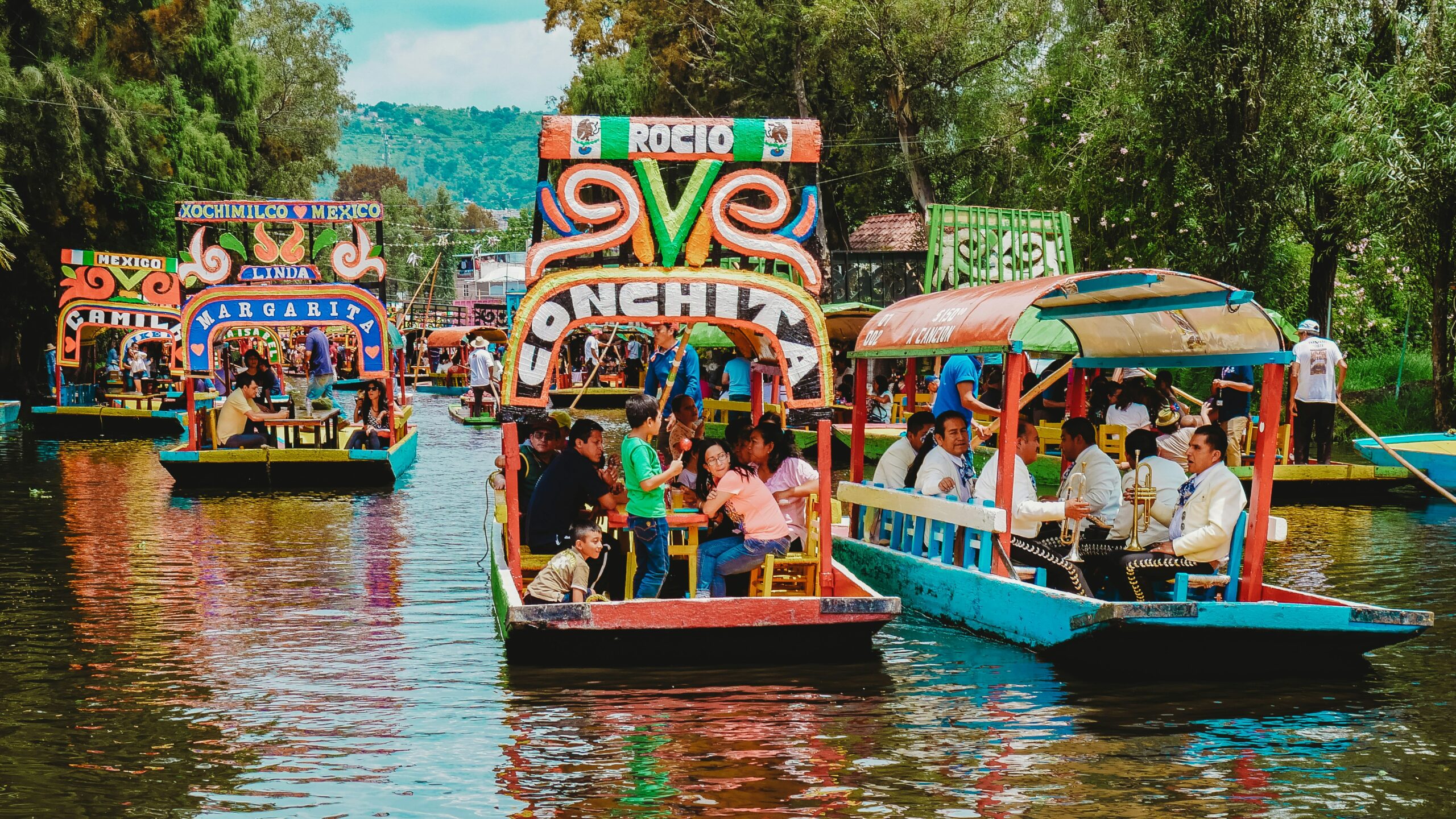 Colorful boats filled with people float on a tree-lined canal in Xochimilco, Mexico, during the day.