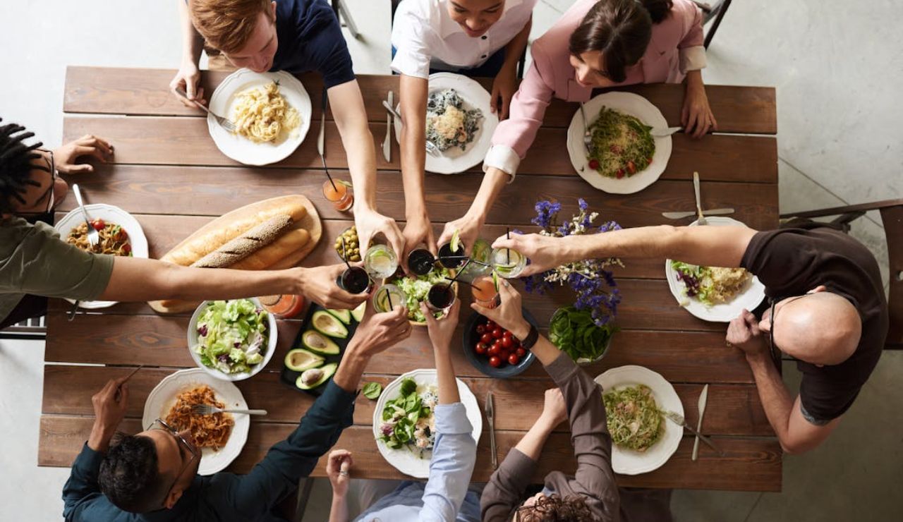 A group of people seated around a wooden table raise their drinks for a toast, surrounded by plates of pasta, bread, salad, and fresh vegetables.
