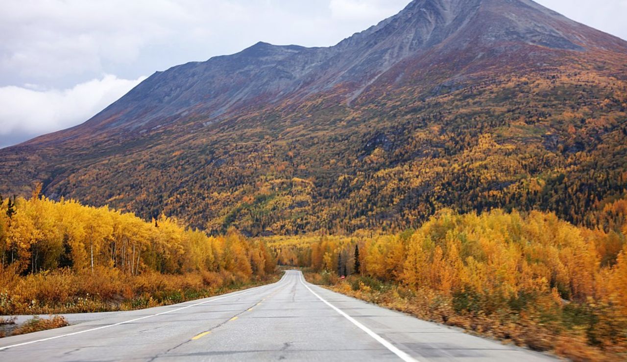 Scenic mountain road lined with vibrant autumn foliage, leading toward a towering, rugged peak under a cloudy sky.