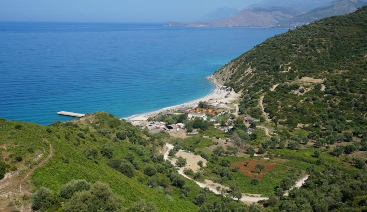 Coastal village nestled between green hills with a beach and clear blue sea stretching into the distance.