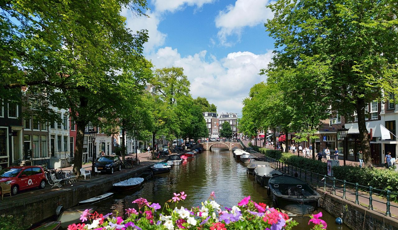 Scenic canal in Amsterdam lined with boats, trees, and traditional Dutch buildings under a sunny sky.