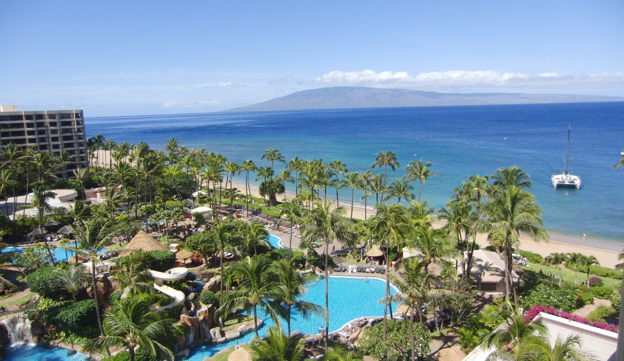 Aerial view of a tropical resort features multiple pools, palm trees, a sandy beach, an anchored boat, and distant islands under a clear sky.