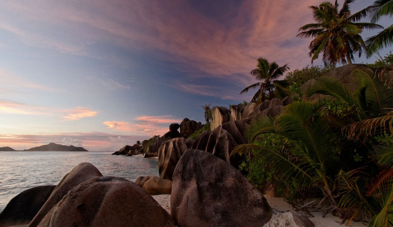 Granite boulders and palm trees lining the shore at Anse Lazio in Seychelles, with a colorful sunset sky over the tranquil ocean.