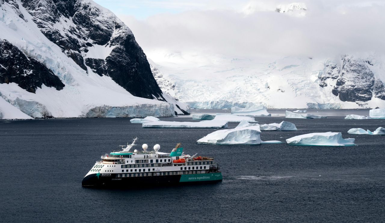 A small icebreaker ship sailing past glaciers and icebergs under a cold sky.