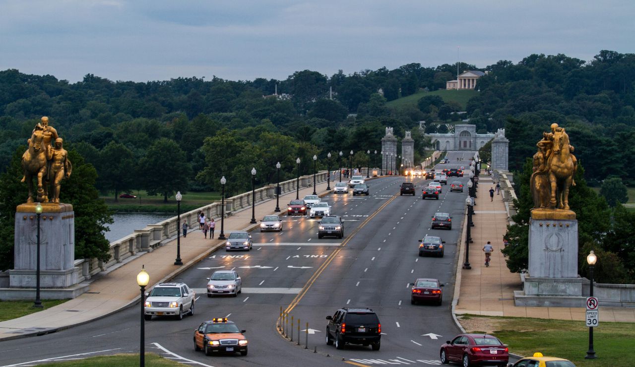 Evening view of Arlington Memorial Bridge with traffic and pedestrians, flanked by golden statues and leading toward the Arlington National Cemetery in the distance.