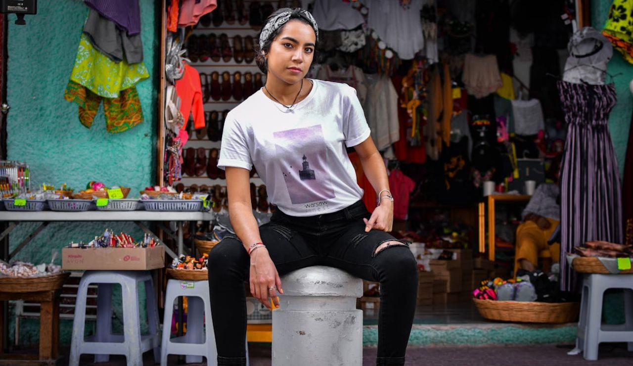 Young woman sitting confidently on a stool in front of a colorful market stall selling clothes and accessories.