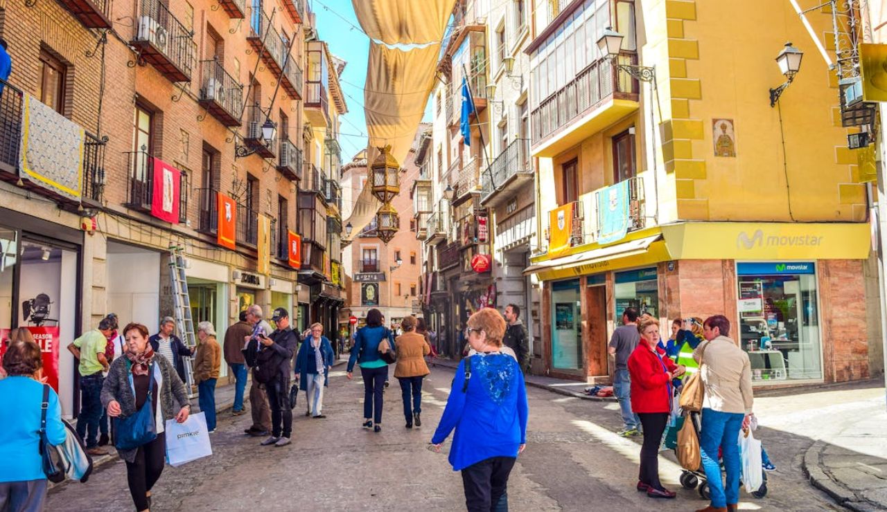 Busy pedestrian street in a European town, with colorful buildings, hanging banners, and people shopping and walking.