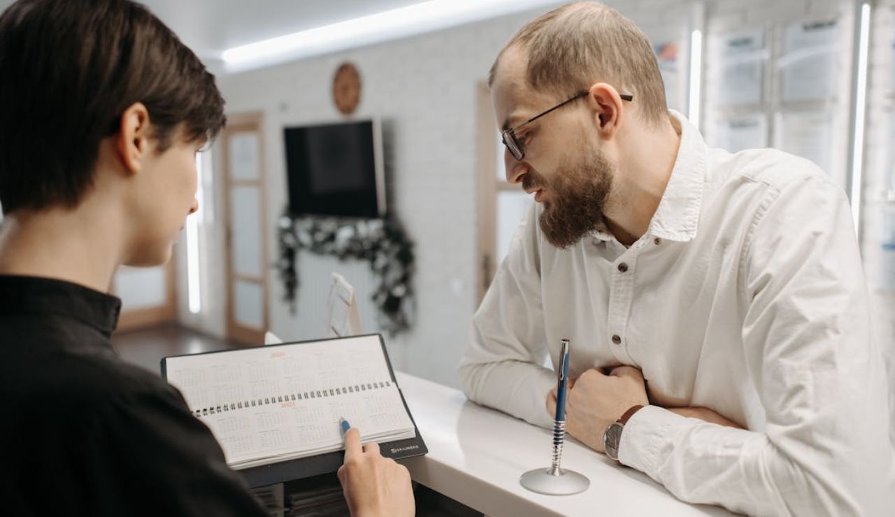 Man at reception desk asking questions while staff shows him a calendar.