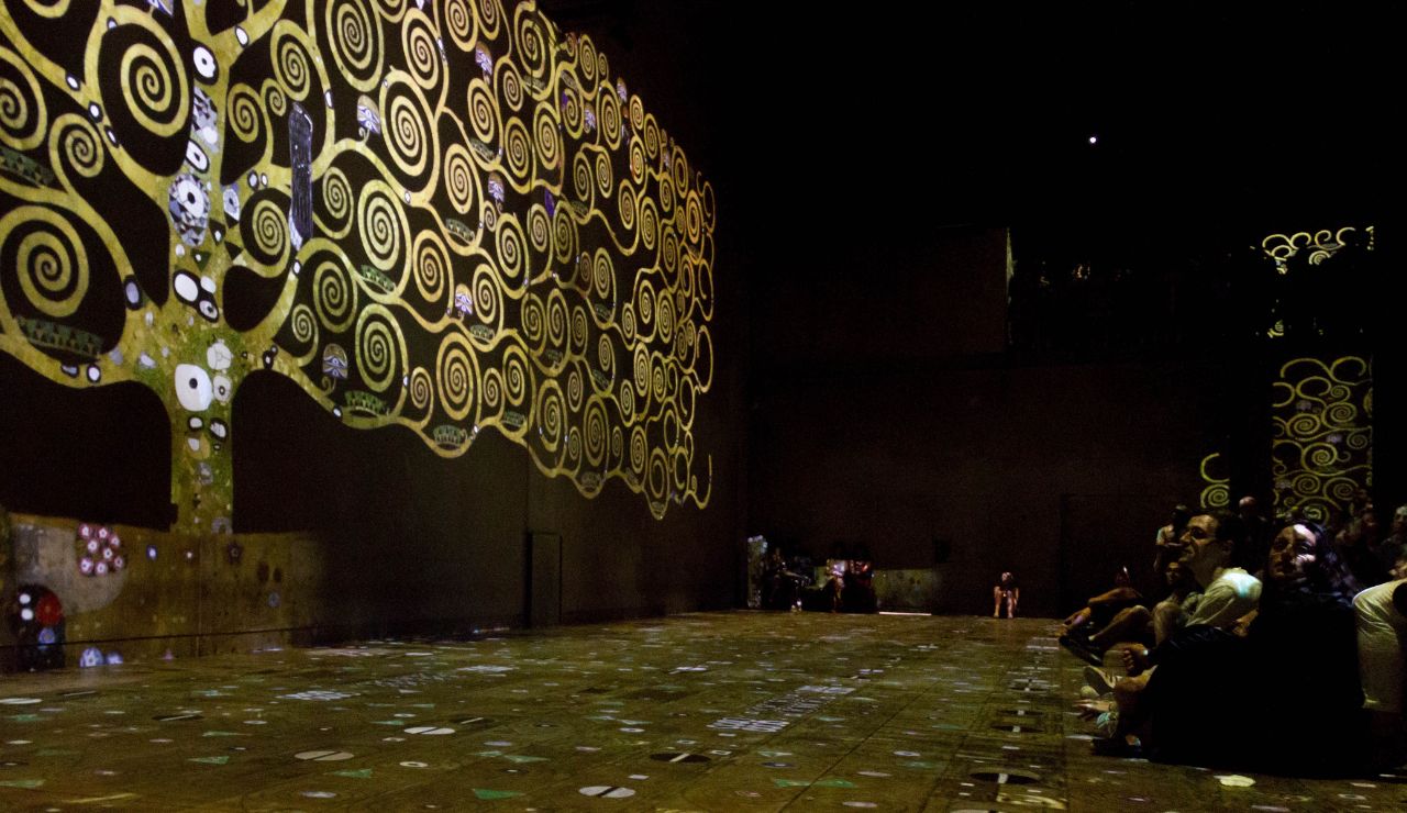 A large group sits in a dark room facing an illuminated wall with a swirling golden tree art installation.